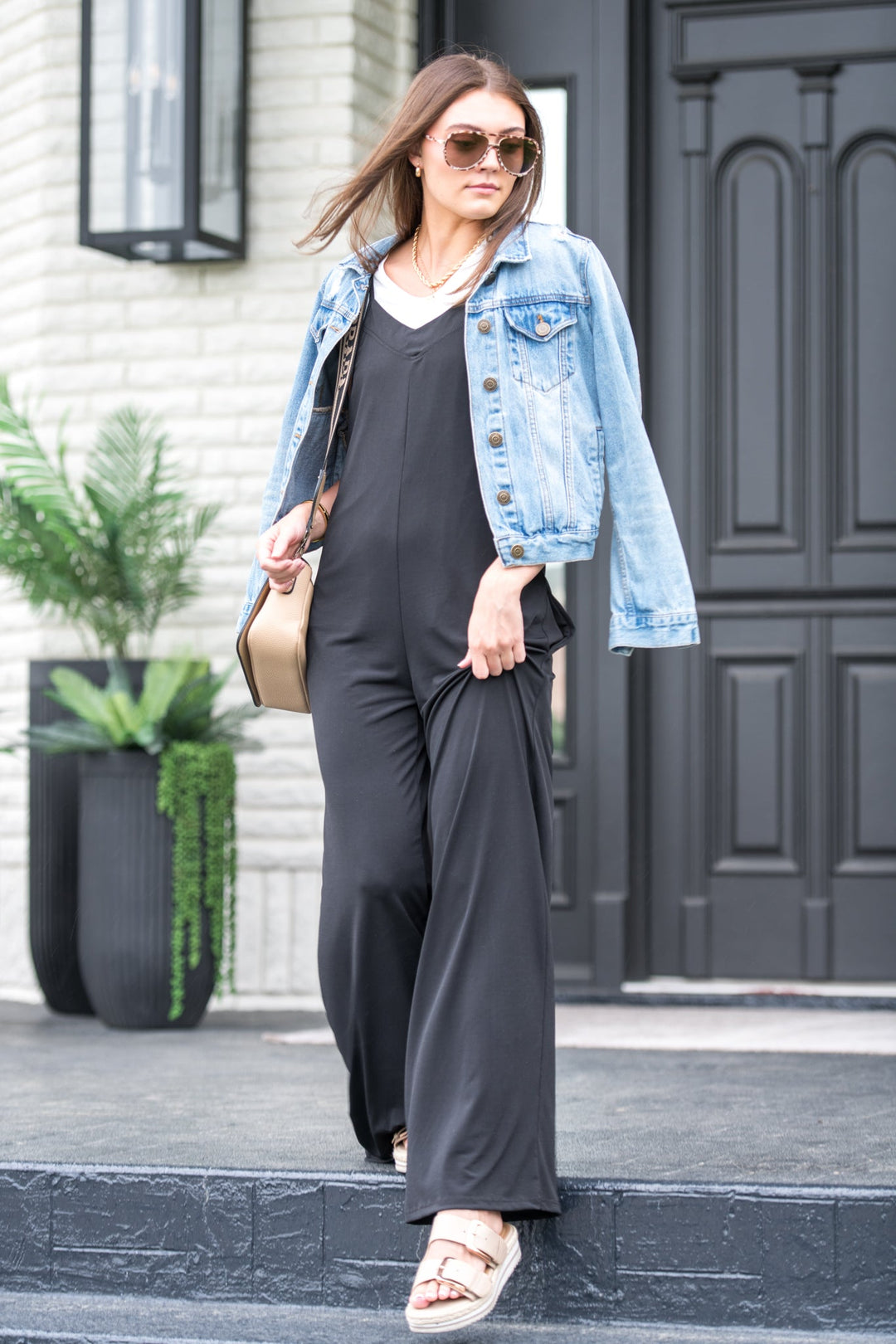 A woman wearing a black tank jumper with a denim jacket, paired with white sandals and carrying a beige bag.