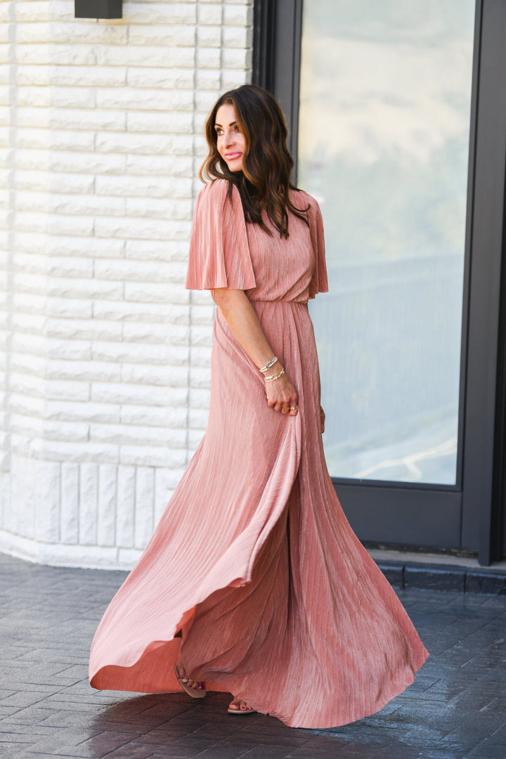 A woman wearing a pink maxi dress with a V-neck and bell sleeves, standing in a flowing pose.