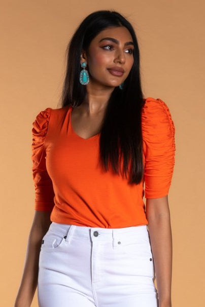 A female model wearing an orange v-neck top with ruched sleeves and white straight-leg pants.
